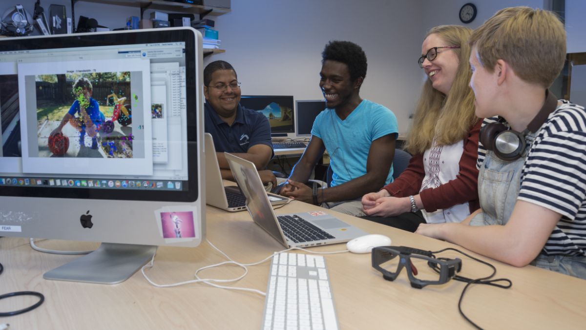 A group of students collaborating around laptops, working on a visual project on screen.