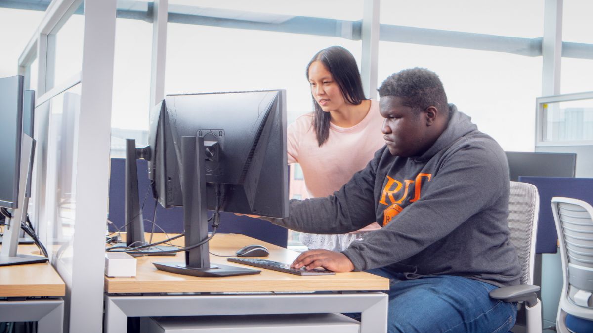 A student seated at a desk with a monitor, receiving guidance from another student standing beside them.