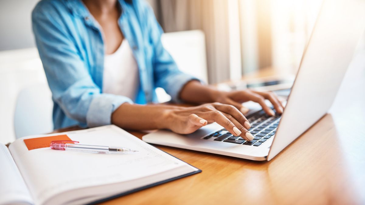 Person typing on a laptop with a notebook and pen on the table under natural sunlight.