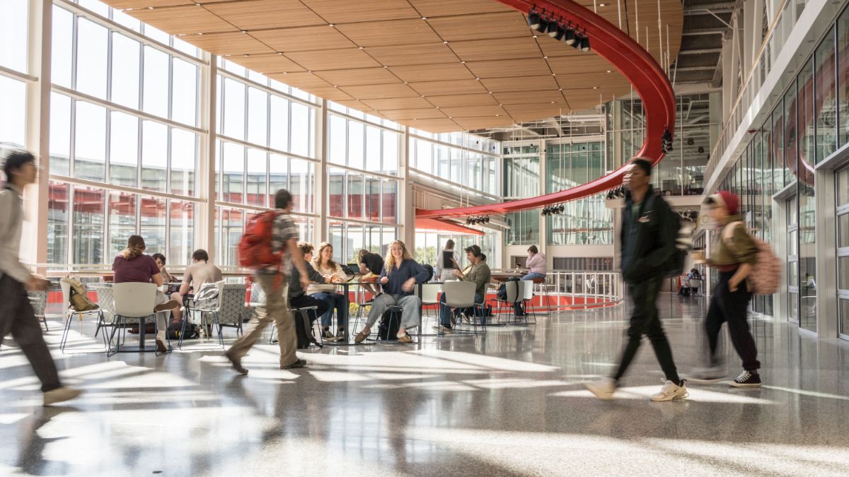 A bright, open atrium with students sitting at tables, while others walk by in motion blur.