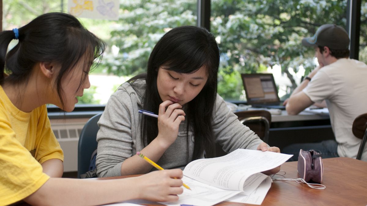 Two students studying together with one assisting the other, sitting by a large window with a laptop in the background.