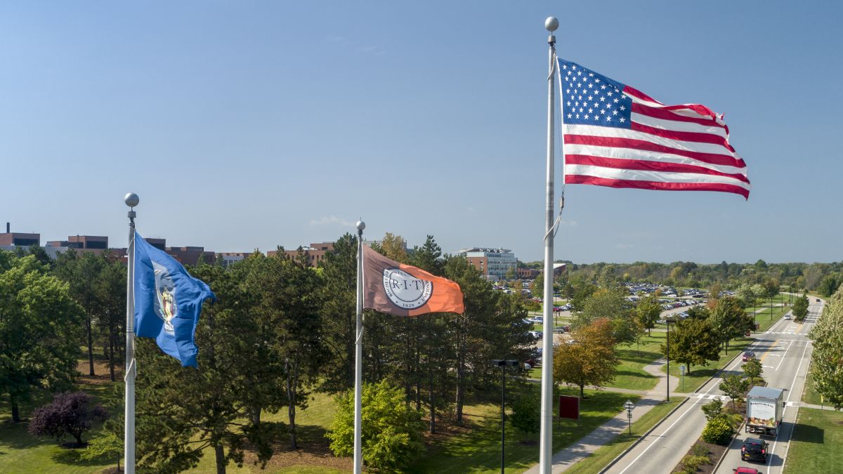 Three flags, including the American flag, the New York state flag, and the R I T flag, flying on a clear sunny day.