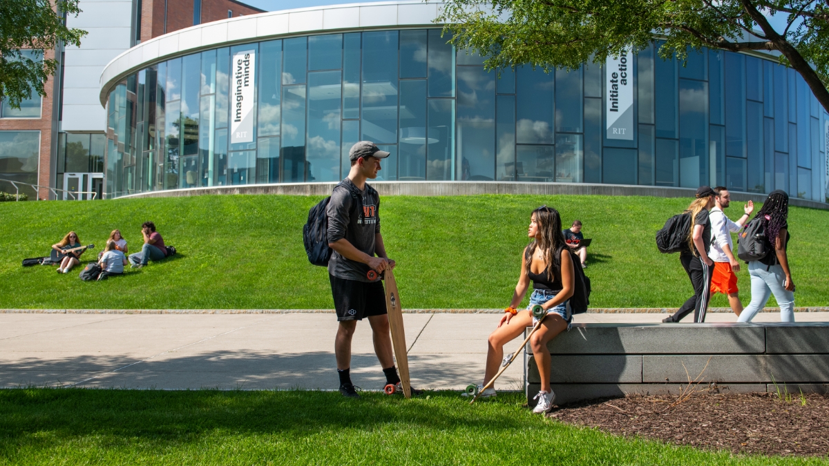 A group of students, some with longboards, chatting and relaxing on a green lawn in front of a modern glass building.