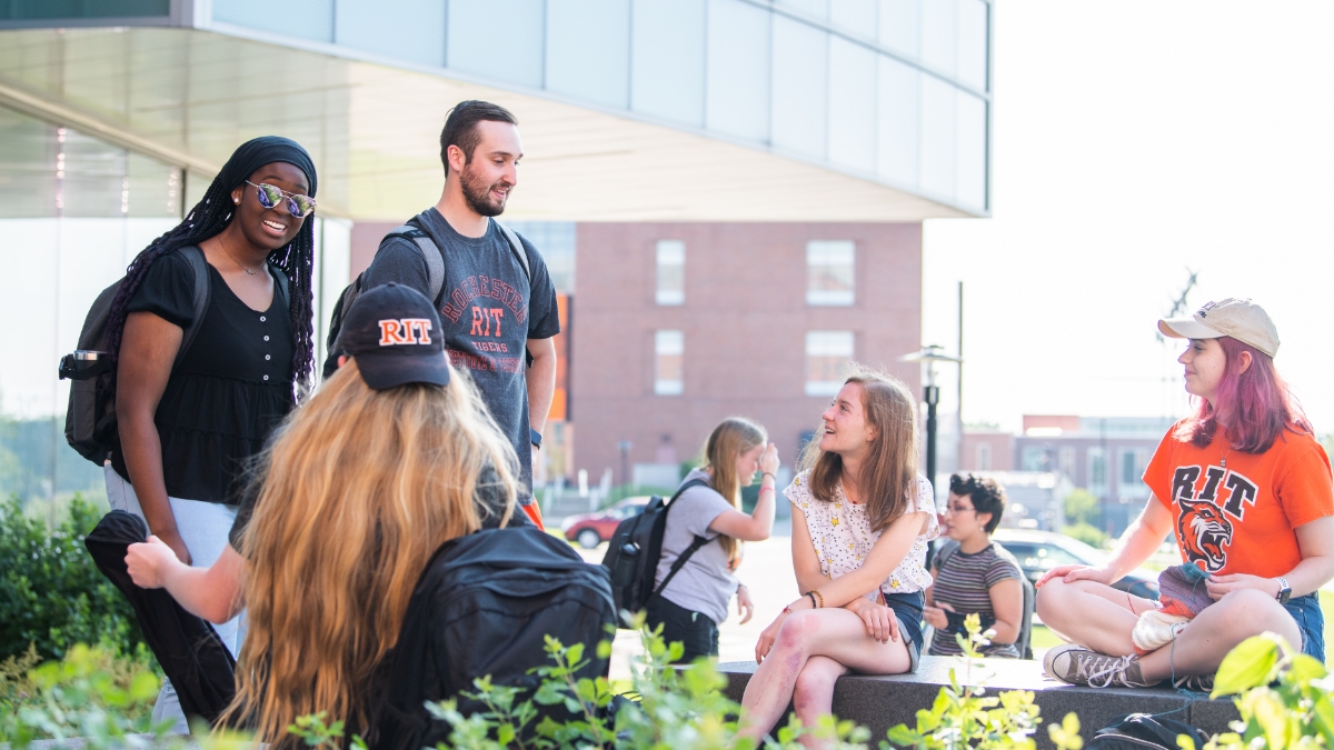 A diverse group of students sits and talks cheerfully outside a campus building.