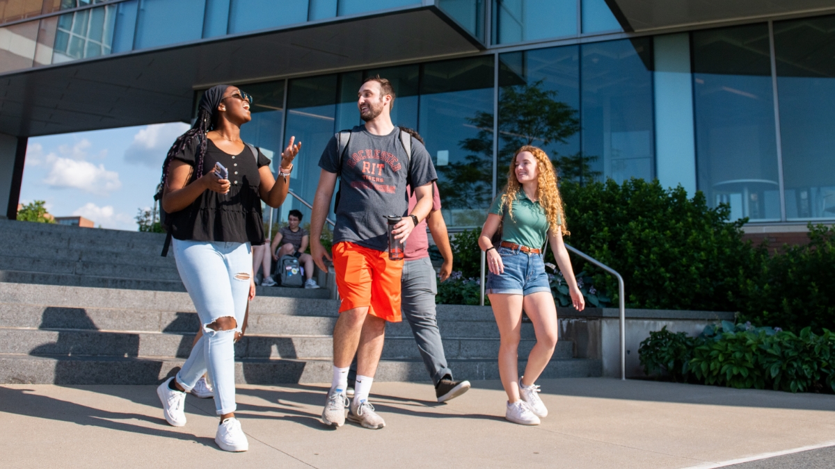 Four students walk down a staircase on campus, talking and laughing as they head towards the path outside a building.