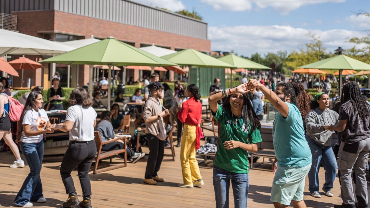 Students socializing and dancing outdoors under green umbrellas during a campus event.