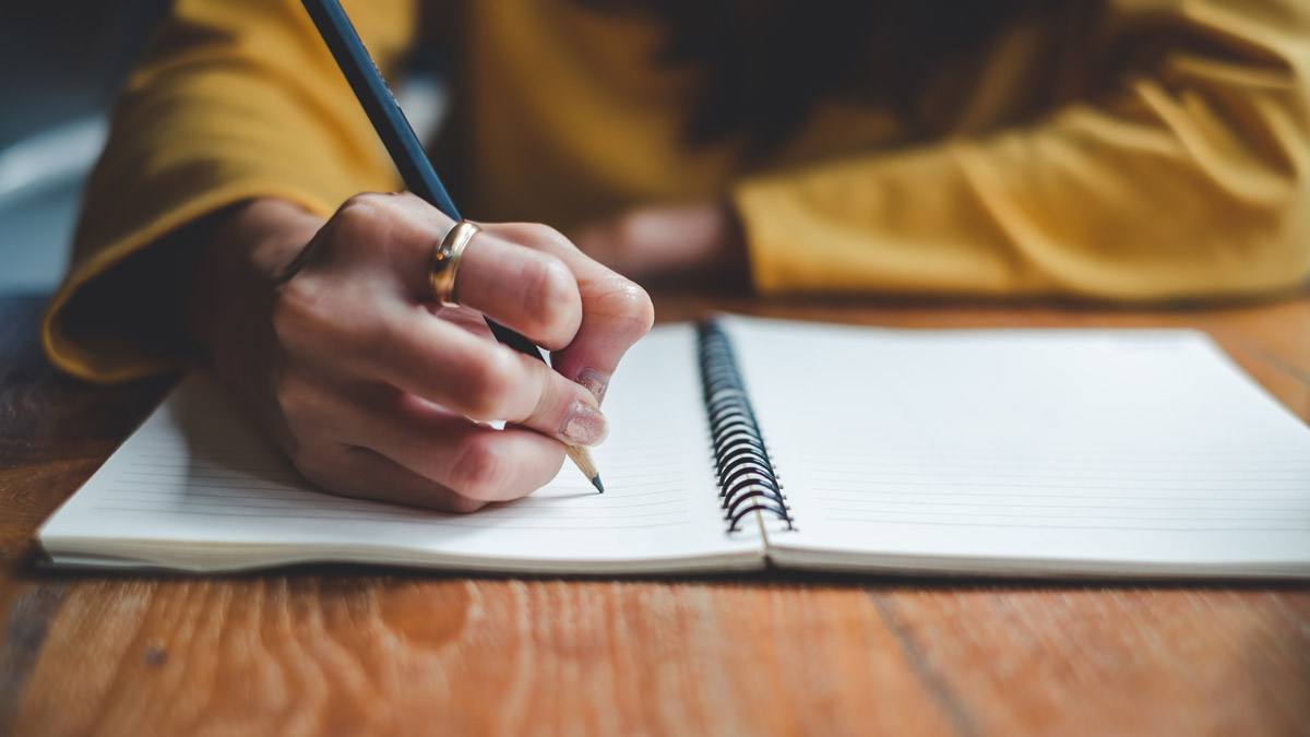 Close up of a hand wearing a gold ring writing in a notebook with a pencil.