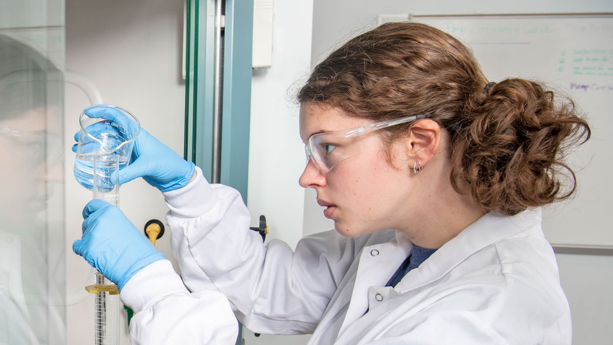 A student in a lab pours clear liquid into a beaker.