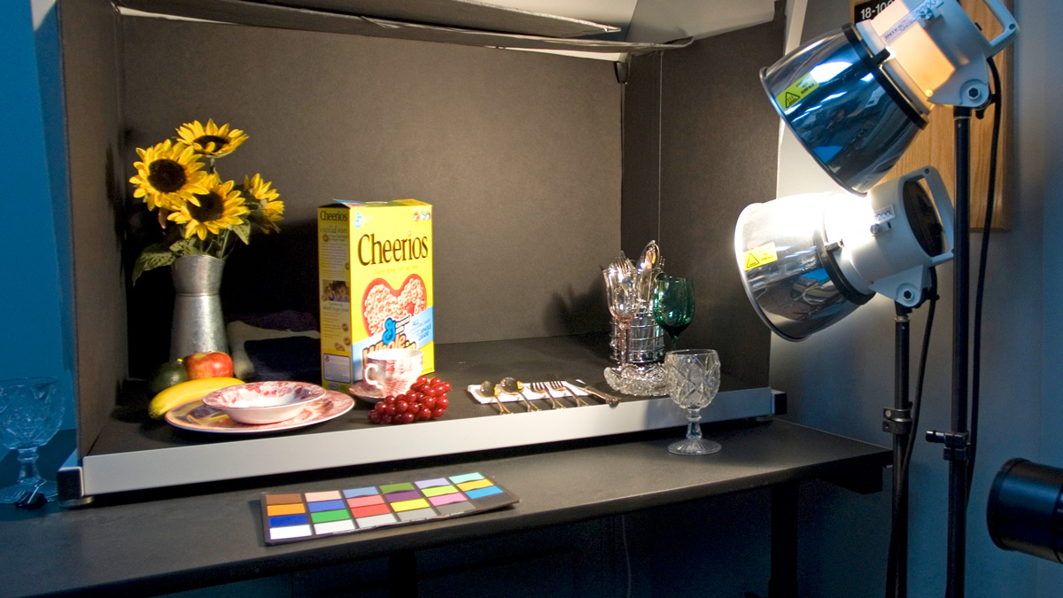 Desk with spotlight shining on Cheerios cereal, plates, sunflowers in vase, utensils, and glasses.