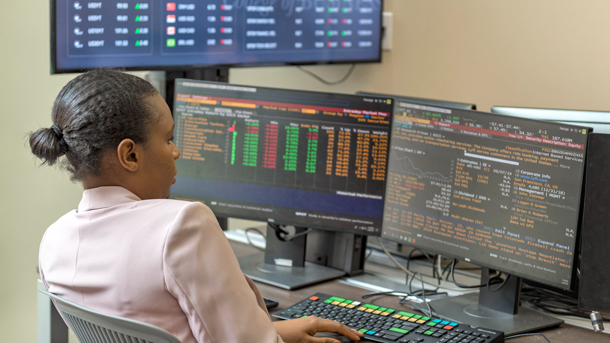 A student sits in front of multiple monitors with various tabular data on them.