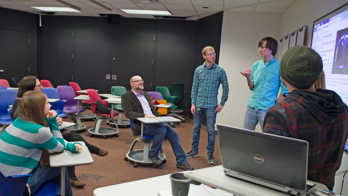 Several people in a classroom are listening to two students standing in front of a projection screen.