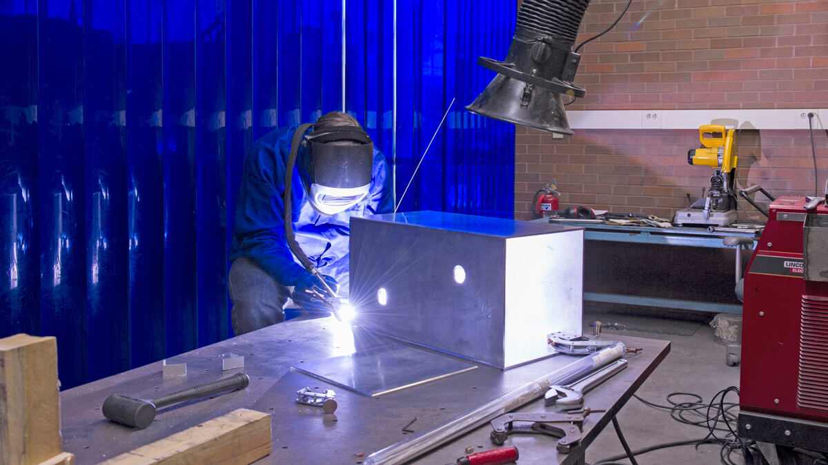 A person wearing a welding helmet welds a metal box.