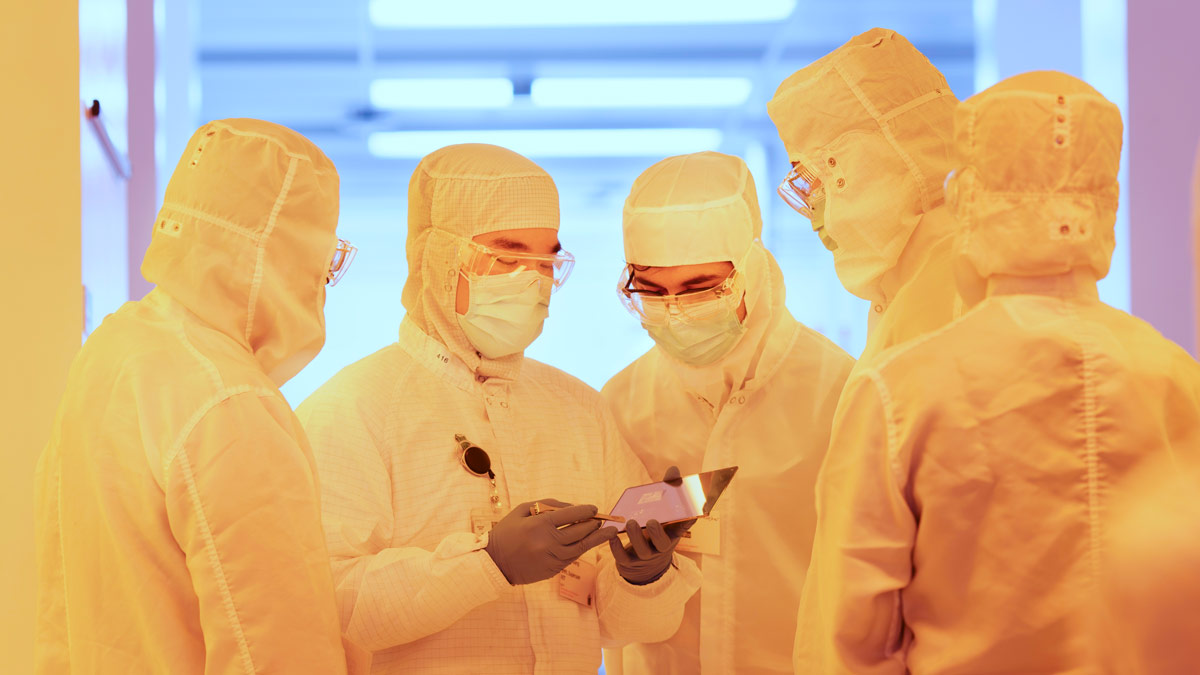 Several people in protective suits in a yellow room look at a computer tablet.