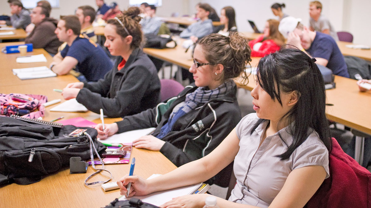 Students at long desks look toward the front of a classroom.