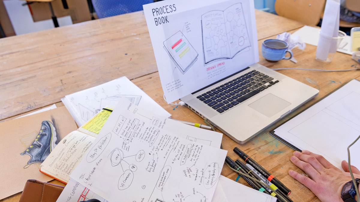 Close up of a laptop, notebook, and several pieces of paper with writing and diagrams.