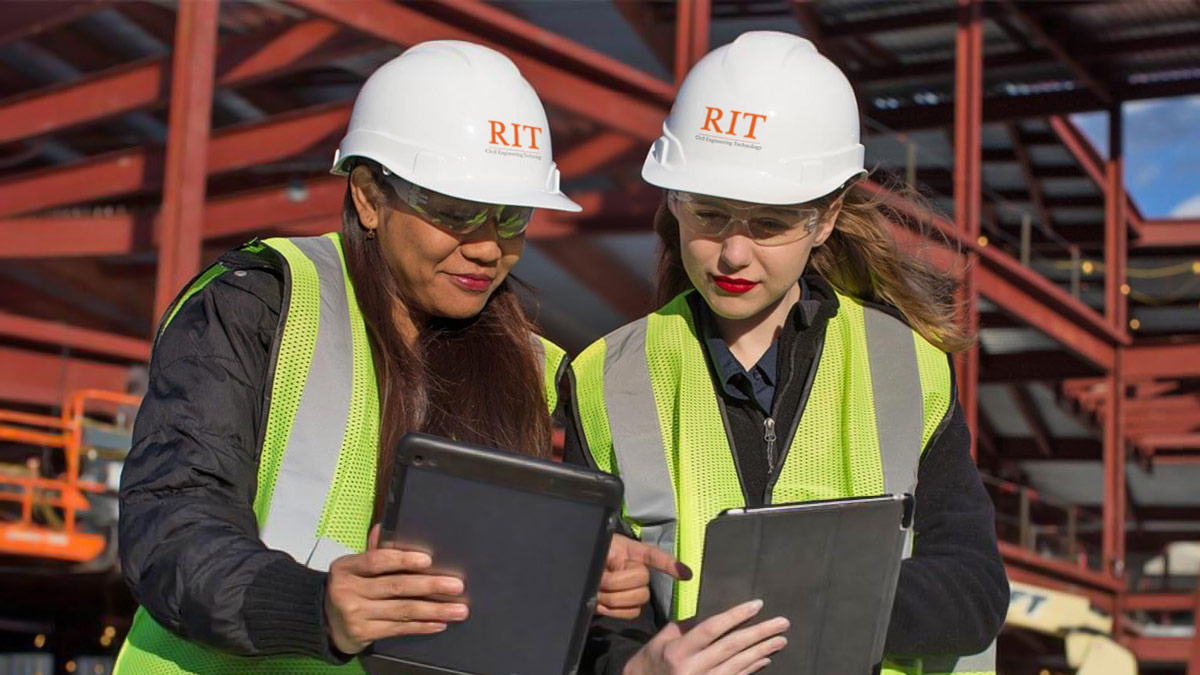 Two people wearing hard hats that say RIT look down at tablet computers. They are standing in from of a construction site.