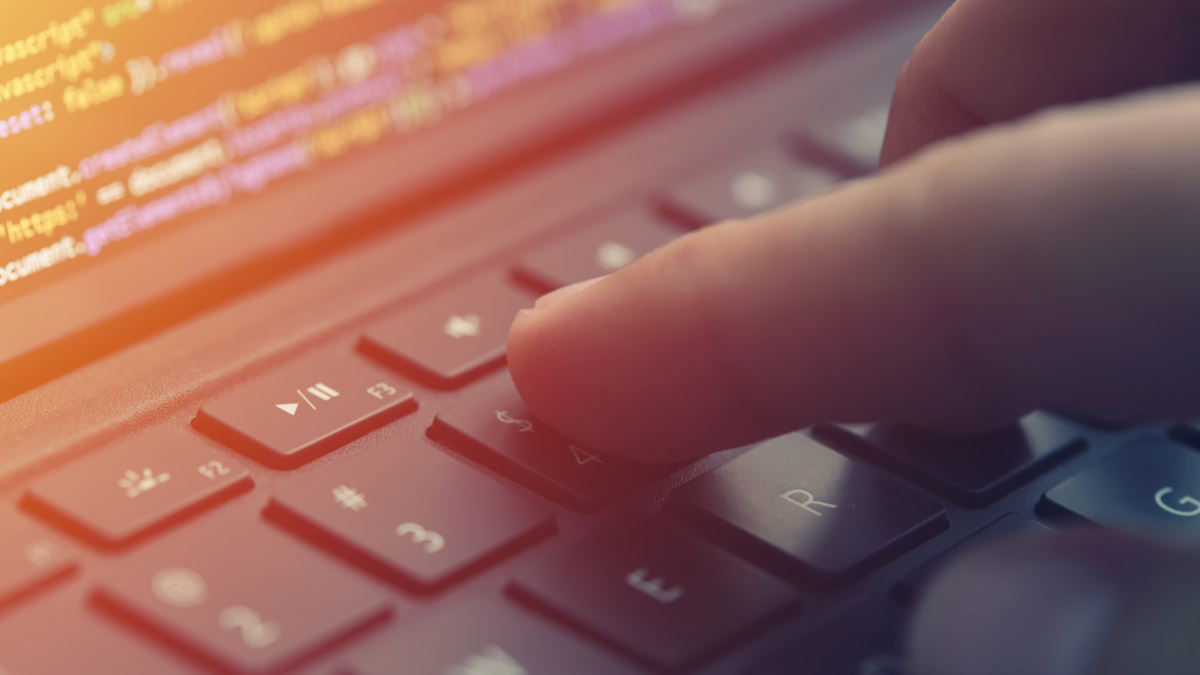 Extreme close up of a finger pressing a key on a laptop keyboard with code visible on the screen.