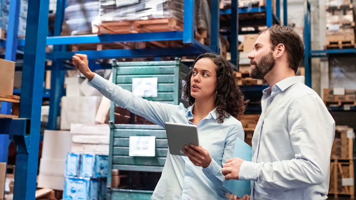 Two people stand in a warehouse looking at stocked shelves and a tablet computer.