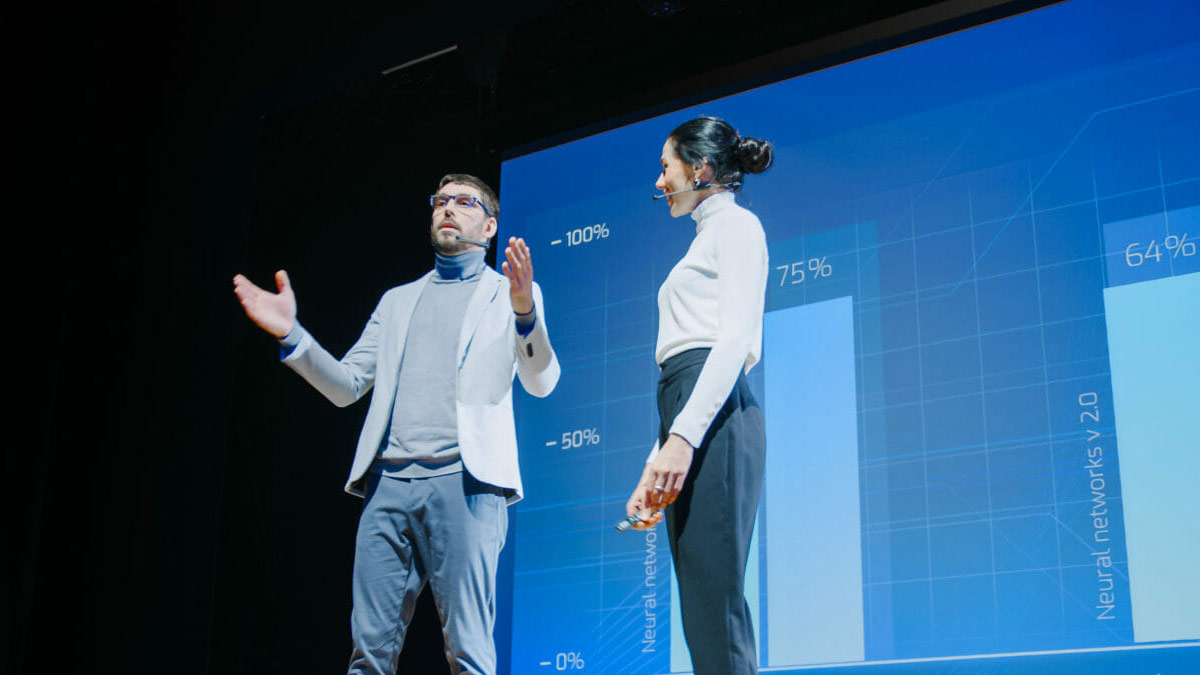 A man and woman stand on stage with charts projected behind them.