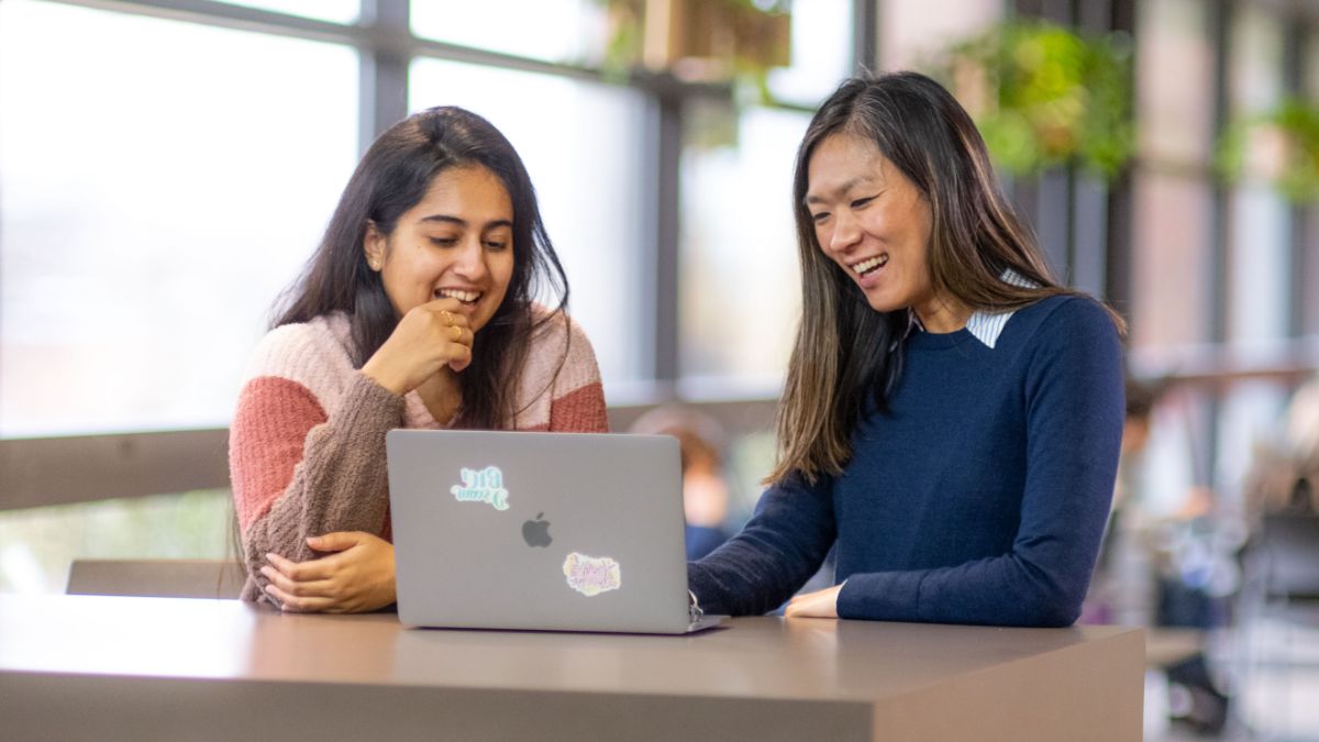 Professor helping a student at a laptop in a lounge or lab.