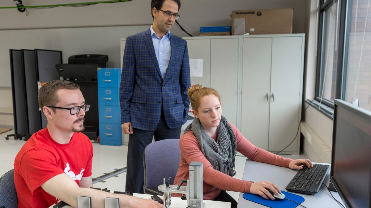 A student sits at a computer, while another has their arm in some measurement device, and a professor stands behind them.