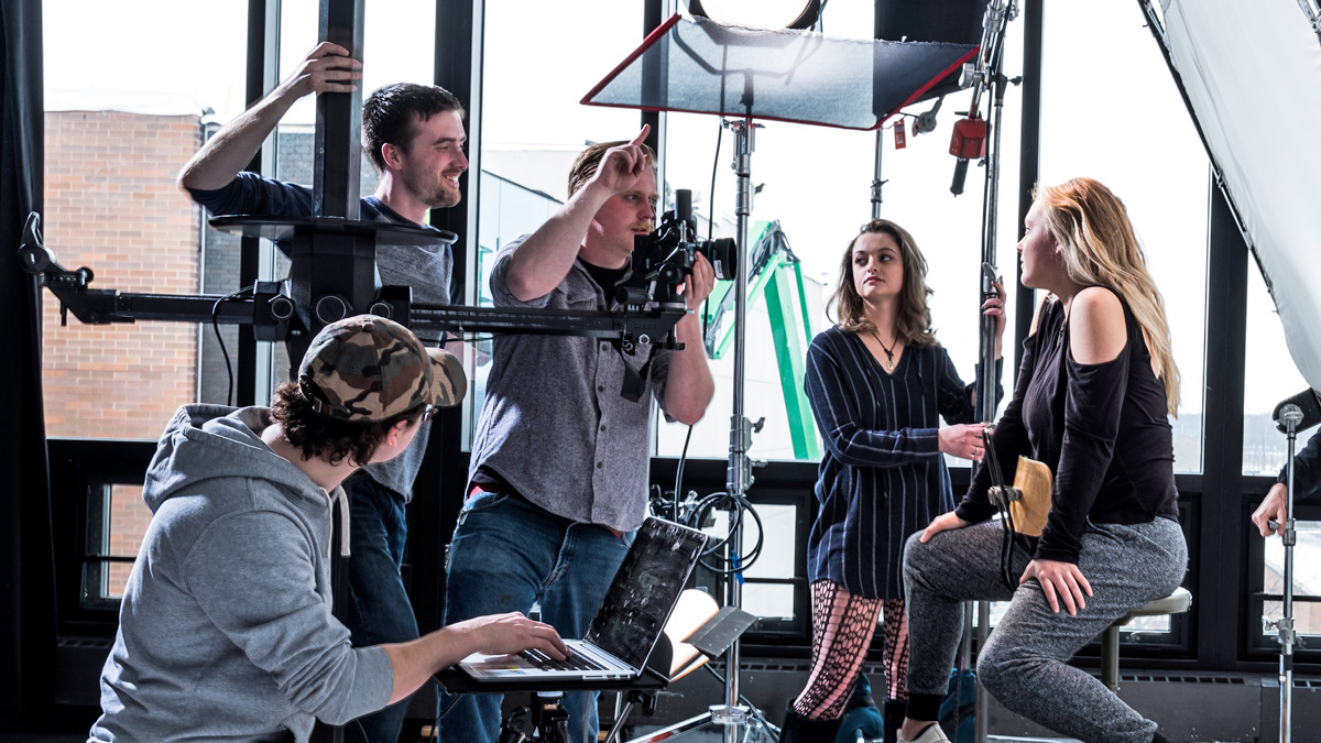 Several students surrounded by studio lights photograph a blond student posing.