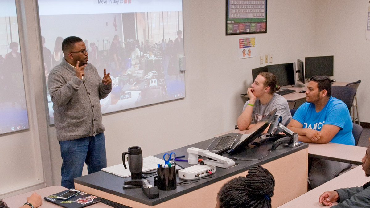 Professor standing in front of whiteboard talking to students in a classroom.