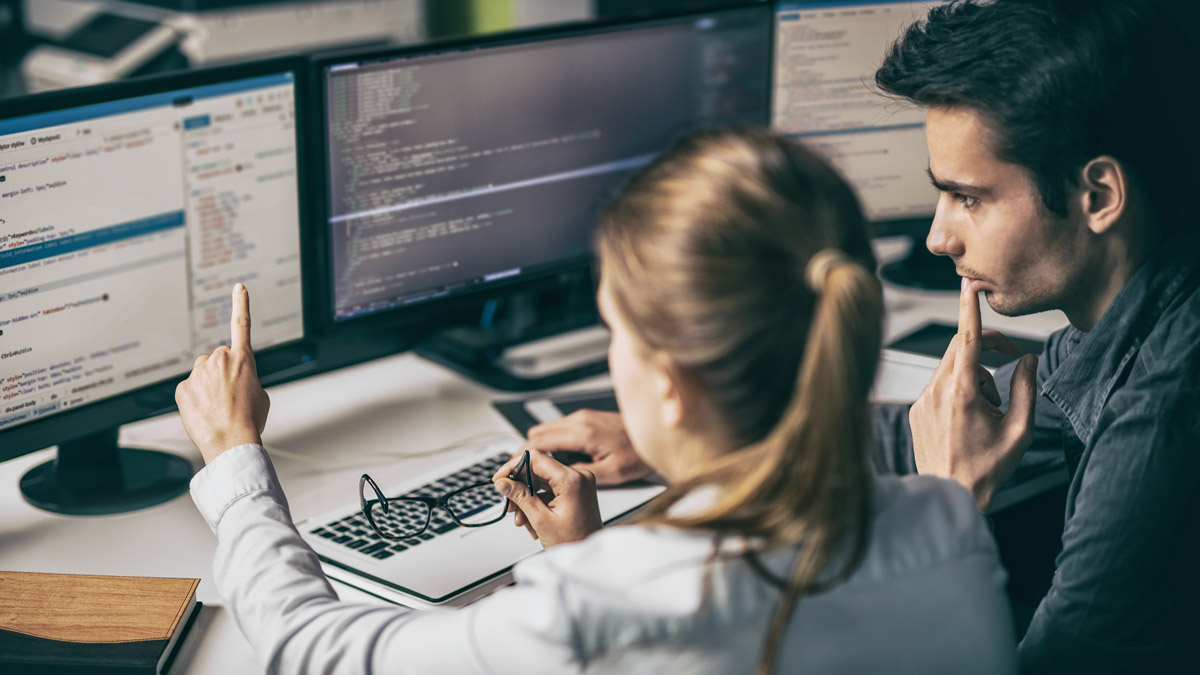 Male and female students sitting at desk with three computer screens displaying code.