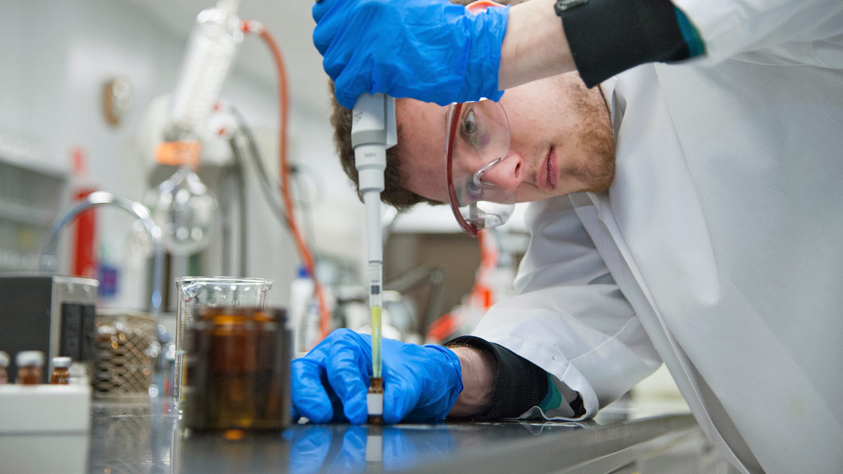 A student wearing a lab coat and protective goggles holds uses a tool to do something outside the crop of the photo.