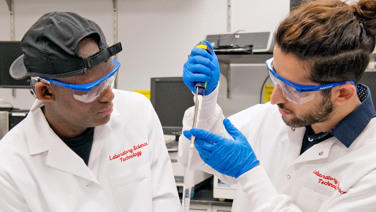 Two students wearing lab coats and protective goggles look at liquid in a pipette.