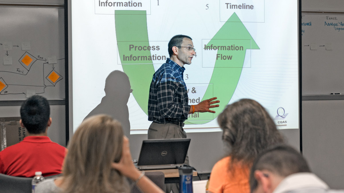 A professor walking in front of projection screen with the text Process Information and Information Flow visible. The backs of student heads are in the foreground.