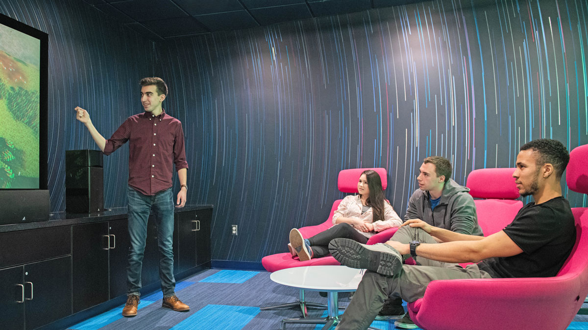 Students sit in a lounge while another standing student points to a projection screen.