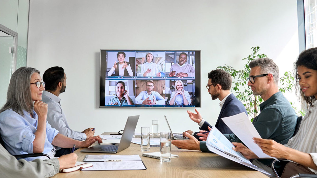 People sit in a conference room with more people joining the meeting remotely, shown on a TV in the center.