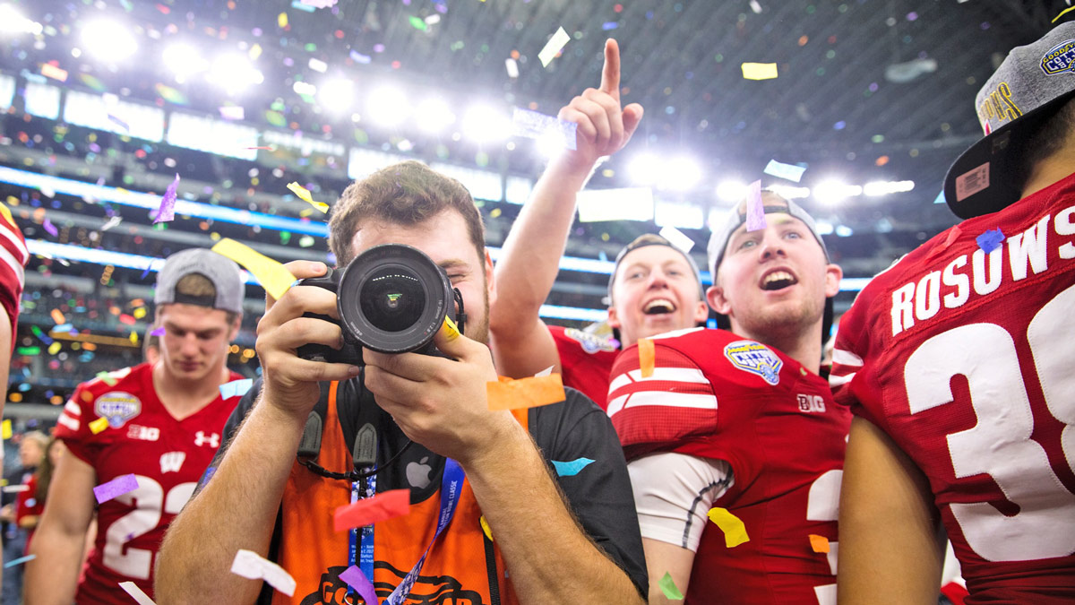 Football players in red stand behind a photographer who's camera is looking straight ahead at the viewer.