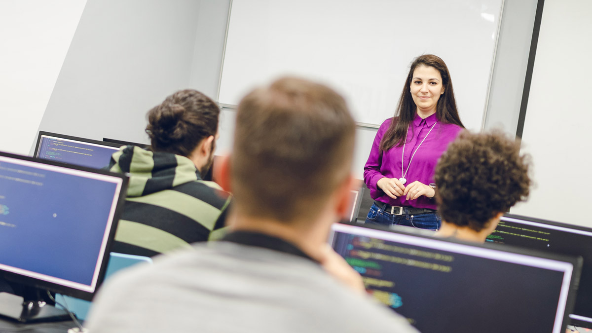 Instructor teaching a class of students sitting at computers with code on screen.