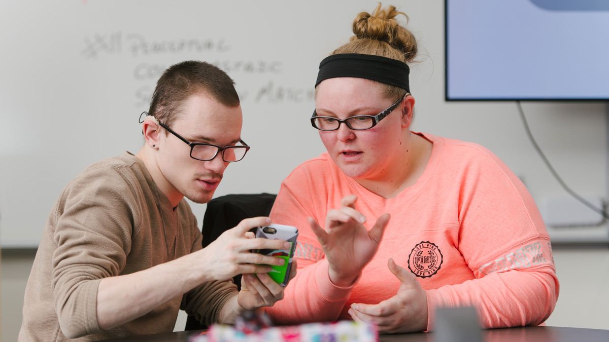 Two students, both wearing glasses, look at a smartphone.