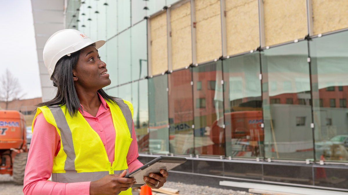 A student wearing a hard hat holds a tablet computer and looks at a construction site with many glass windows.