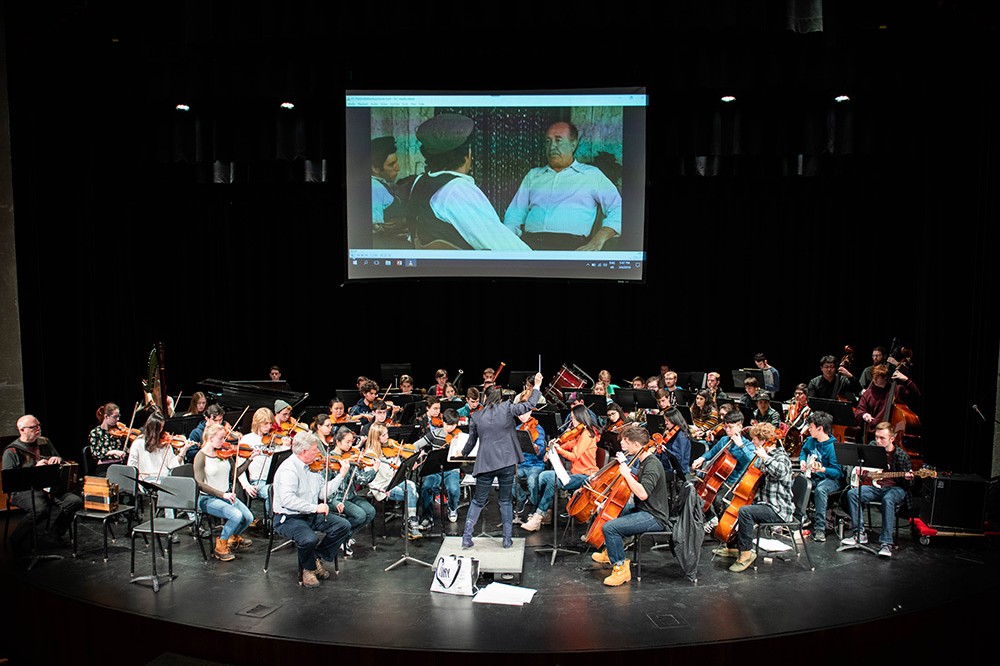 orchestra performs on stage with screen showing a movie behind them