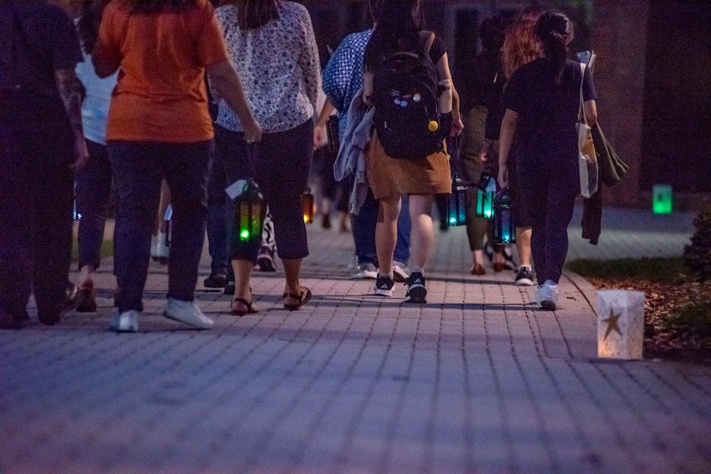 people are shown from the waist down walking away from the camera with small white illuminated bags lighting a pathway next to them.