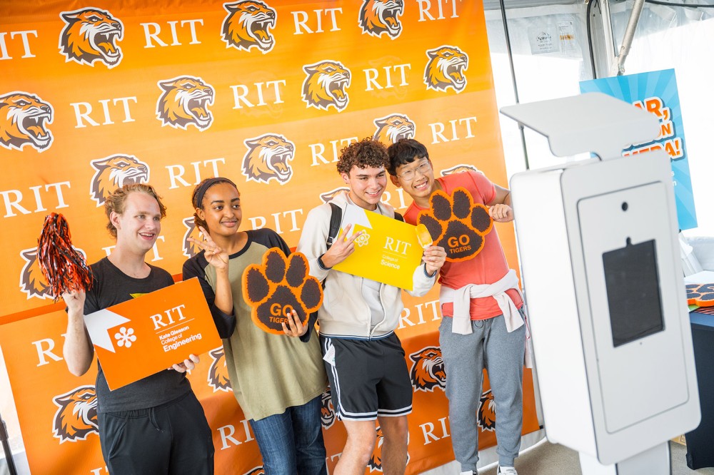 Four students stand in front of an R I T branded step and repeat with orange and black tiger accessories for a photo.