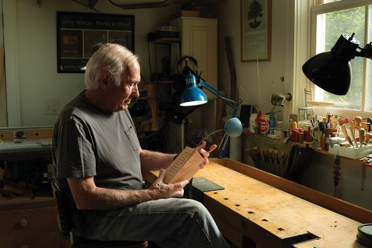 man in a woodworking shop seated at a workbench holding a rectangular piece of wood.