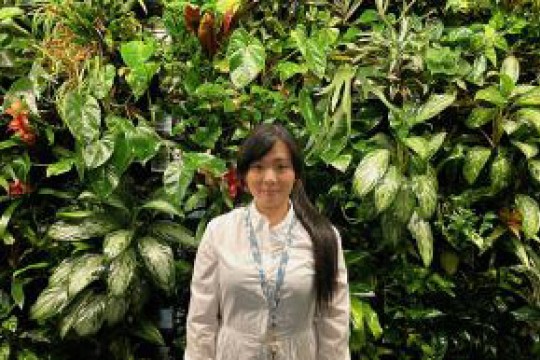 ' Hui-yu Ho stands in a cream colored blazer in front of a botanical plant wall.'