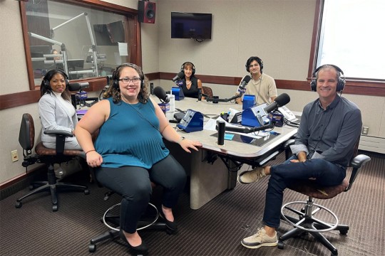 'Five people are shown sitting around a table in a beige room wearing headphones.'