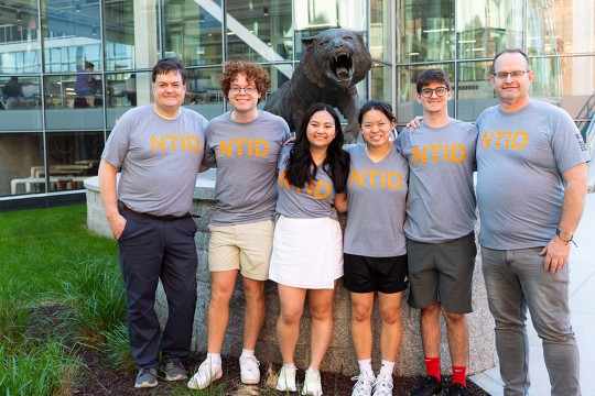 a group of six people wearing R I T shirts stand in front of the SHED and a large cement tiger on R I T's campus