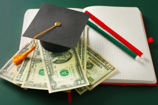 a graduation cap sits on top of american bills of various value which sits atop an open notebook with 2 colored pens on the corner.