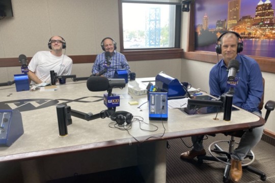 Three men wearing headphones sit at a table in a radio talk studio: the man on the left is bald and is wearing glasses and a white t-shirt; the man in the center is wearing a blue and white plaid long sleeved button down shirt; the man on the right has short dark hair and is wearing a blue long sleeved button down shirt and jeans.