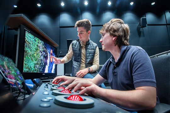 'two students stand in front of a small screen reviewing footage in the MAGIC Center Color Correction Suite on campus.'