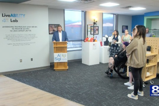 a man stands at a poduim next to a wall that reads LiveABILITY lab. Members of the media stand around him as he speaks.