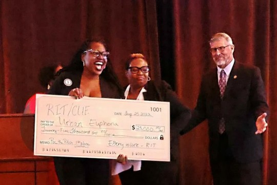 2 women of color stand with a giant size check for $25,000. A man stands next to them with his arms extended in celebration.