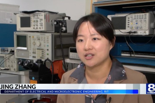 Jing Zhang wears a brown blazer and sits in a semiconductor lab speaking to W R O C TV.